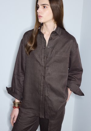 Person mit langen braunen Haaren trägt ein lockeres dunkelbraunes Hemd mit Knöpfen und passende Hose, ergänzt durch goldene Ohrringe und Armbänder.