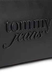 Εμβαπτισμένο λογότυπο "tommy jeans" σε μαύρο χρώμα σε υφή μαύρου δέρματος, με γυαλιστερό φινίρισμα στα ανάγλυφα γράμματα.