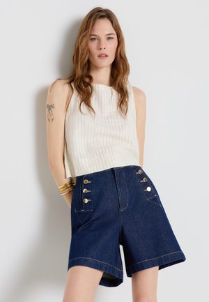 Donna con capelli castani mossi in piedi contro un muro bianco, indossa una canotta bianca a righe sottili senza maniche e pantaloncini di jeans scuri a vita alta con bottoni dorati.