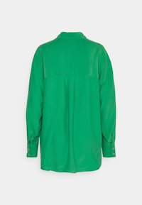 Chemise verte à manches longues avec poignets boutonnés, vue de dos sur un fond clair uni.