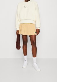 Krémová fleecová mikina, béžová plisovaná sukně s malým vyšívaným detailem, hnědá chlupatá kabelka, bílé ponožky a tenisky.