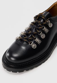 Schwarzer Lederschuh mit dicker Sohle, gelbgestreifte schwarze Schnürsenkel, die durch dekorative silberne Haken und Ösen gefädelt sind.