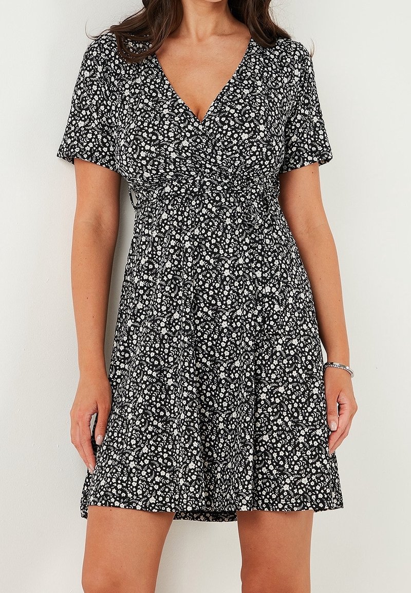 Femme portant une robe courte noire avec un motif floral blanc, col en V et manches courtes, debout devant un mur blanc uni.