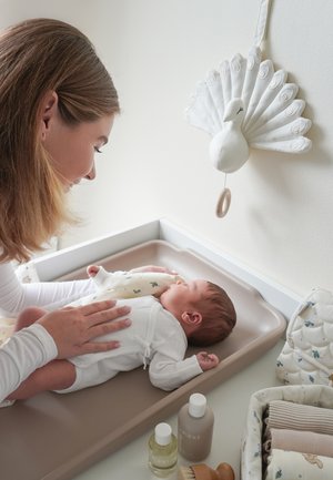 Mor smiler og rører forsigtigt ved nyfødt, der ligger på puslemåtte med hvid påfuglevægdekoration og babyplejeartikler i nærheden.