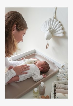 Madre sorridente che tocca delicatamente il neonato sdraiato sul fasciatoio con una decorazione da parete di pavone bianco e articoli per la cura del bambino nelle vicinanze.