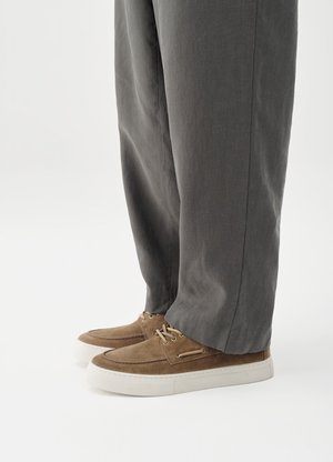 Personne portant des chaussures à lacets en daim marron et un pantalon gris à jambes droites, debout sur un fond blanc.
