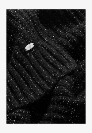 Fekete kötött anyag bordás textúrával, fehér fémes szálakkal, és egy apró ezüst fém címkével, amely jól láthatóan kiemelkedik.