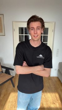 Νεαρός άνδρας με ανοιχτό καστανά μαλλιά στέκεται με σταυρωμένα τα χέρια, φορώντας μαύρο μπλουζάκι με λογότυπο και ανοιχτόχρωμο τζιν σε φωτεινό μοντέρνο δωμάτιο.