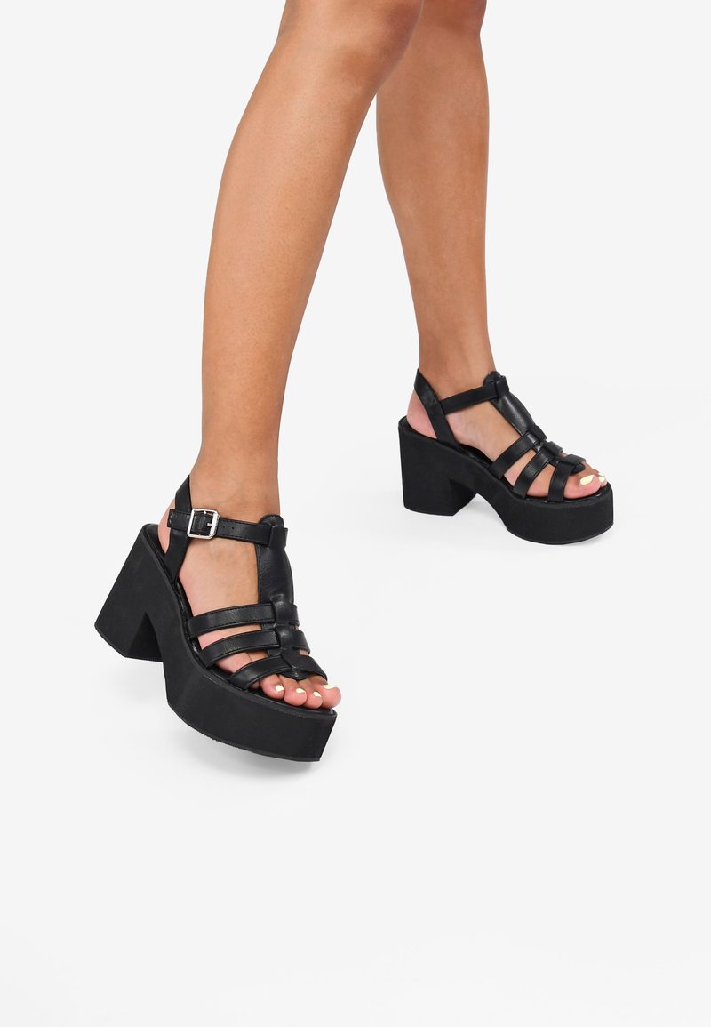 Sandalias de plataforma negras con suelas gruesas, diseño de tiras y cierre de hebilla. Textura de cuero suave con punta abierta.