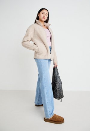 Beige fleece jacket with high collar, light pink top, light blue flared jeans, and brown slip-on shoes. Holding a dark patterned bag.