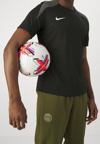 Černé sportovní tričko se šedými akcenty, v kombinaci s olivově zelenými kalhotami s zlatým logem. Vícebarevný fotbalový míč je držen pod paží.