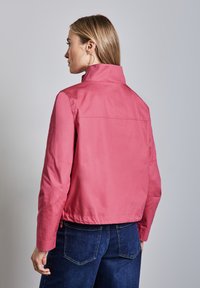Veste courte rose avec un col montant et des manches longues. Présente une texture lisse et un design légèrement court. Portée avec un pantalon en denim.