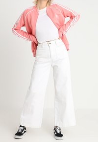 Rosa zip-up träningsjacka med vita ränder, vit åtsittande topp, högmidjade vita vida jeans och svarta sneakers.