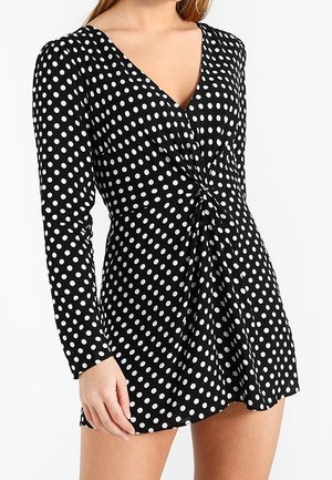Musta mekko, jossa on pitkät hihat ja syvä V-pääntie, vyötäröllä solmukohta ja valkoisia pilkkuja kauttaaltaan.