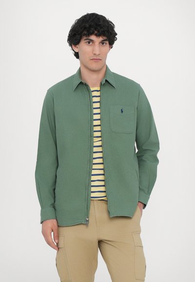 Camisa verde con cremallera, hecha de tela tejida, que cuenta con un bolsillo en el pecho y un logo sutil, usada sobre una camiseta a rayas amarilla y azul marino.