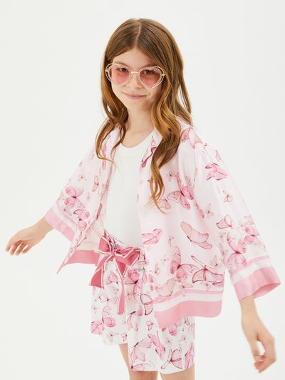 Completo rosa chiaro con stampe di farfalla, con un nastro coordinato in vita. Comprende una maglietta ampia e pantaloni corti, completati da occhiali da sole a forma di cuore.