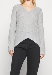 Pull en maille gris clair avec un col en V, des manches longues et un design à ourlet asymétrique. Présente un tissu texturé et des poignets ajustés.