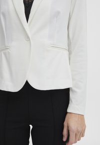 Biely blazer so priliehajúcim strihom, ktorý obsahuje dve predné vrecká, jedno gombíkové zapínanie a hladkú textúru, skombinovaný s čiernymi nohavicami.