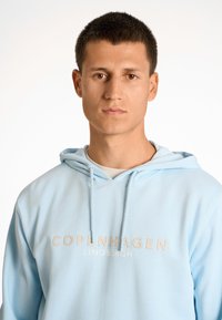 Sudadera azul claro hecha de tela suave, con cordones y texto impreso "COPENHAGUE LINDBERGH" en colores contrastantes en la parte frontal.