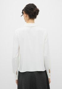 Camicia bianca a maniche lunghe con una fenditura laterale sui polsini, dotata di colletto classico e texture liscia, abbinata a una gonna a righe. 