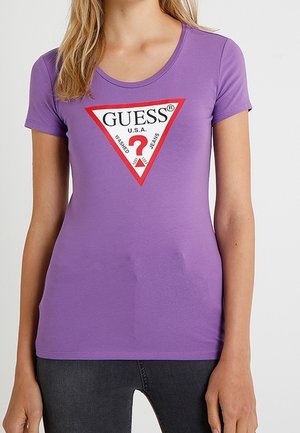 Kvinde iført en tætsiddende lilla T-shirt med et rødt og hvidt trekantet Guess-logo og sorte jeans, stående mod en ensartet lys baggrund.