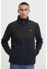 Giacca in pile blu navy con un pannello nero sul petto, colletto alto e chiusura con zip; presenta tasche laterali e dettagli con logo.