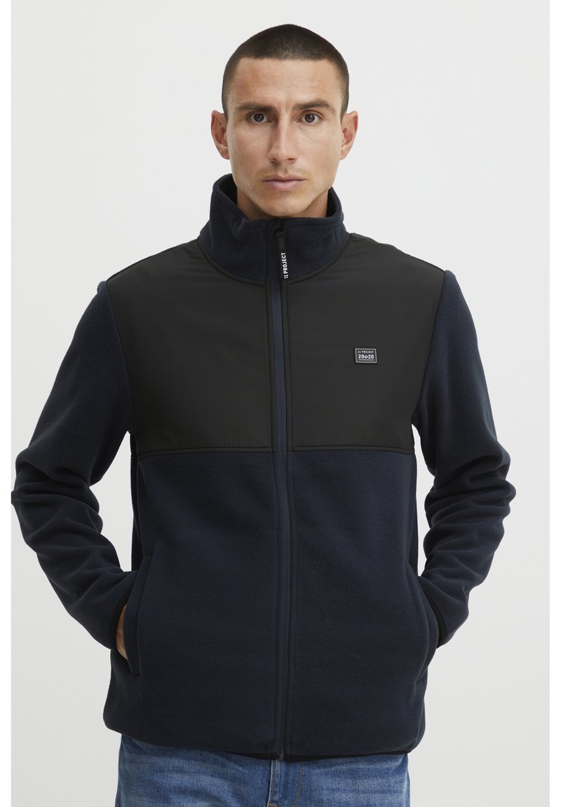 Giacca in pile blu navy con un pannello nero sul petto, colletto alto e chiusura con zip; presenta tasche laterali e dettagli con logo.