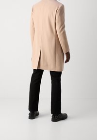 Abrigo de lana rosa claro con corte recto, abertura en la espalda y mangas largas, combinado con pantalones negros ajustados y botines negros.
