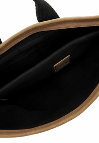 Bolsa de tela abierta en color bronceado y negro que muestra un interior espacioso negro con un bolsillo interior con cremallera y un parche de cuero cosido con el logo.