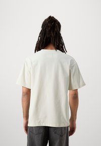 Ljusbeige kortärmad t-shirt med rund halsringning och en rektangulär etikettdetalj på baksidan, tillverkad av mjuk bomullstyg.
