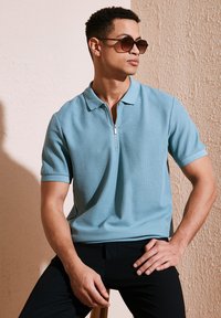 Kreklu ar īsām piedurknēm un ar rāvējslēdzēju, gaismā zilā krāsā, fakturēts audums. Valkāts ar melniem biksēm. Izcelti saulesbrilles ir tumši brūnas ar noapaļotu formu.