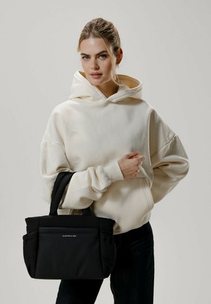 Borsa a tracolla nera con due manici, caratterizzata da un'esterno testurizzato e una tasca frontale. Il modello indossa un hoodie color crema e jeans scuri.