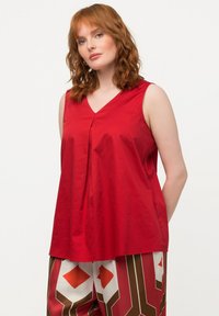 Blusa rossa senza maniche con scollatura a V, caratterizzata da un dettaglio plissé sul davanti. Texture liscia, vestibilità comoda, abbinata a pantaloni con patter.