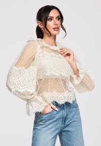 Femme portant un chemisier en dentelle crème transparente à manches bouffantes et un jean bleu, posant avec une main dans la poche et l'autre près du col.