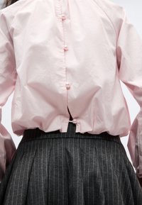 Ανοιχτό ροζ μπλούζα με κουμπιά από μαργαριτάρια στο πίσω μέρος, συνδυασμένη με ψηλόμεση πλισέ φούστα σε σκούρο γκρι με ρίγες.