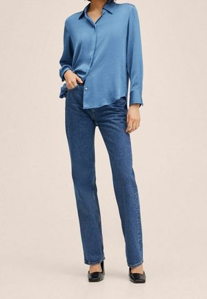 Kobieta w jasno niebieskiej bluzce z długim rękawem, wsadzonej w proste niebieskie dżinsy, w czarnych szpilkach o zaostrzonych czubkach, stojąca na neutralnym tle.