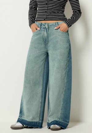 Personne portant un jean en denim bleu bicolore à jambes larges avec des ourlets effilochés et un haut à manches longues rayé noir et blanc, mains dans les poches.