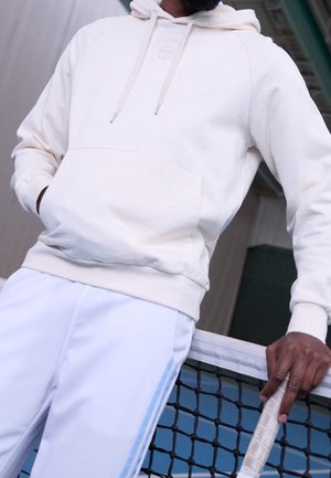 Mężczyzna w białej bluzie z kapturem i białych spodniach, opierający się o siatkę tenisową, trzymający rakietę tenisową z owiniętym uchwytem.