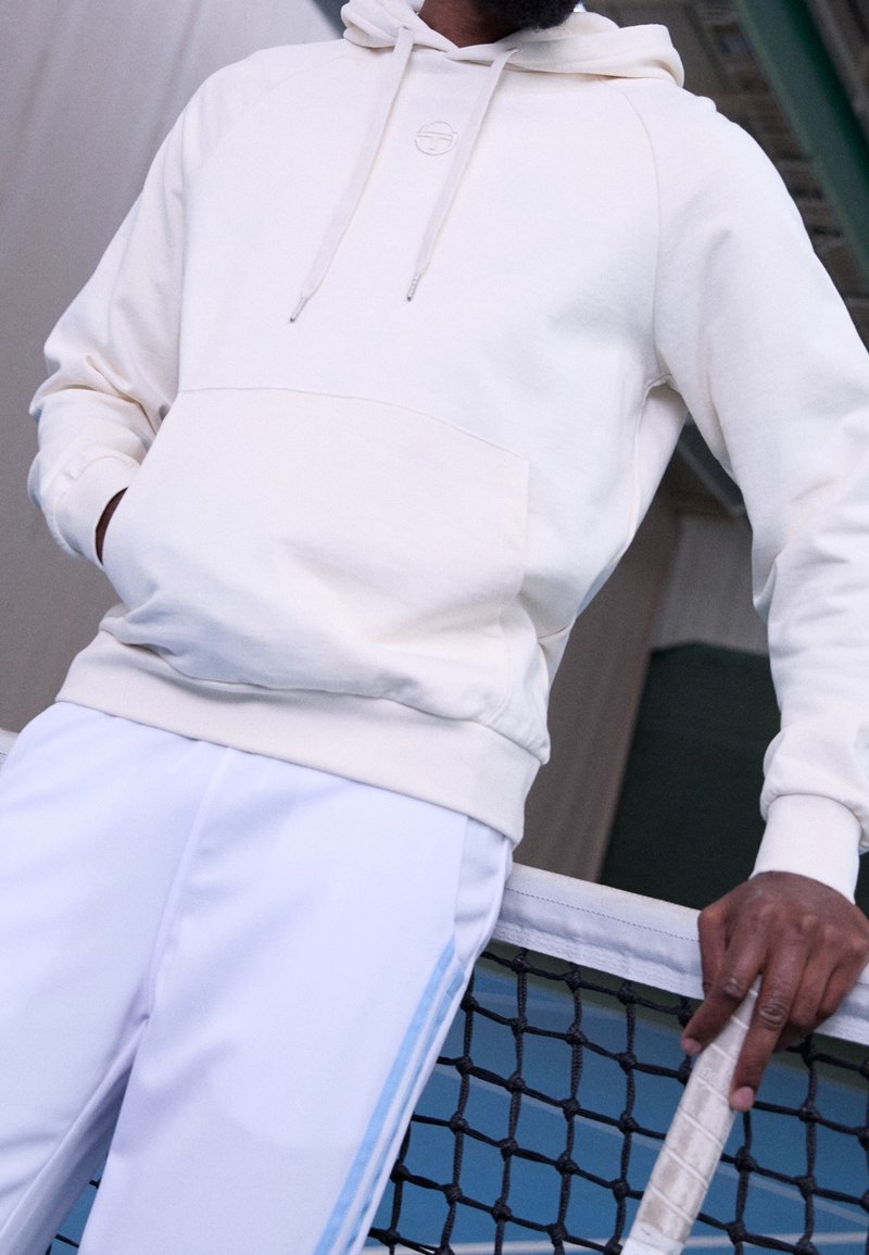 Homme en sweat à capuche blanc et pantalon blanc appuyé sur le filet de tennis, tenant une raquette de tennis avec une poignée bandée.