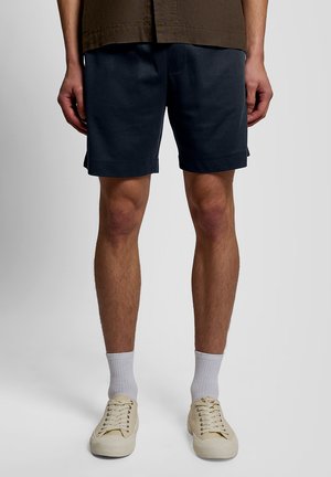 Homme portant un short bleu marine, des chaussettes blanches montantes et des baskets lacées beige clair, debout devant un fond uni gris clair.