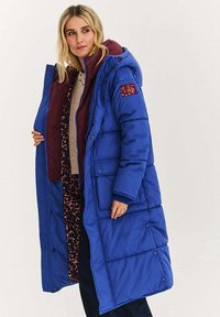 Niebieska puchowa kurtka z bordową podszewką i wnętrzem w panterkę. Posiada kaptur, przednie kieszenie oraz kontrastową łatę na rękawie.