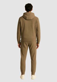 Felpa con cappuccio a coste marrone e joggers abbinati con vestibilità rilassata. Presenta un cappuccio, polsini elasticizzati e gambe slim-fit. Sneakers visibili.
