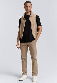 Bež pleten brezrokavnik čez črno majico, združeno s tan hlačami in belimi supergami. Enostavna zasnova brez vidnih vzorcev ali poudarkov.