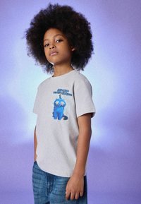 Graues T-Shirt mit kurzen Ärmeln und einem blauen Cartoon-Katzendesign mit großen Augen und Blitzelementen. Kombiniert mit blauen Jeans.