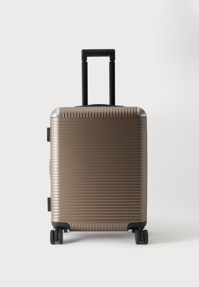 Valise cabine coque dure beige avec des nervures horizontales, poignée télescopique noire déployée, et quatre roues pivotantes sur fond blanc.