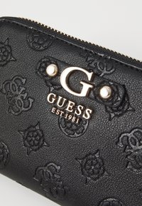 Portofel negru texturat cu modele florale embossate, având un logo GUESS proeminent de culoare aurie și închidere cu fermoar.