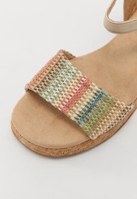 Sandal avec lanières tissées et motif multicolore en rouge, vert et bleu. Dispose d'une semelle en liège beige et d'un coussin de pied en suède doux.