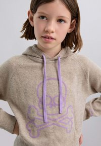 Beige Kapuzenpullover mit weicher Textur, ausgestattet mit einem lila Totenkopf- und Kreuzschnüre-Design. Enthält lavendelfarbene Zugkordeln und gerippte Bündchen.