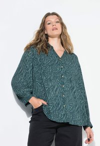 Blusa verde con un motivo a zebre testurizzato, maniche lunghe, apertura con bottoni davanti e una vestibilità morbida, abbinata a pantaloni neri.
