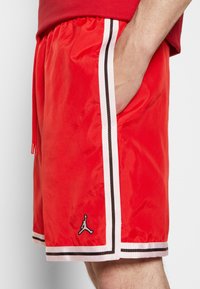 Shorts de sport rouges en tissu léger, avec des rayures latérales blanches et brunes, une taille élastique et un logo sur l'ourlet.